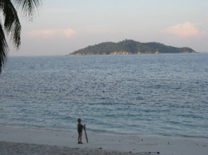 Pieni poika seisoo trooppisen saaren rannassa hiekalla ajopuuhun nojaten ja merelle katsoen.