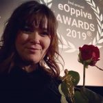 Heli Järvenpää hymyilee palkintopysti ja ruusu kädessä. Taustalla valkokangas, jossa heijastettuna "eOppiva Awards 2019" -tunnus.
