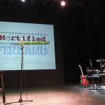 Esiintymislavalla tyhjä penkki, rummut, mikrofoni ja seinälle heijastettuna teksti "Mortified Finland".